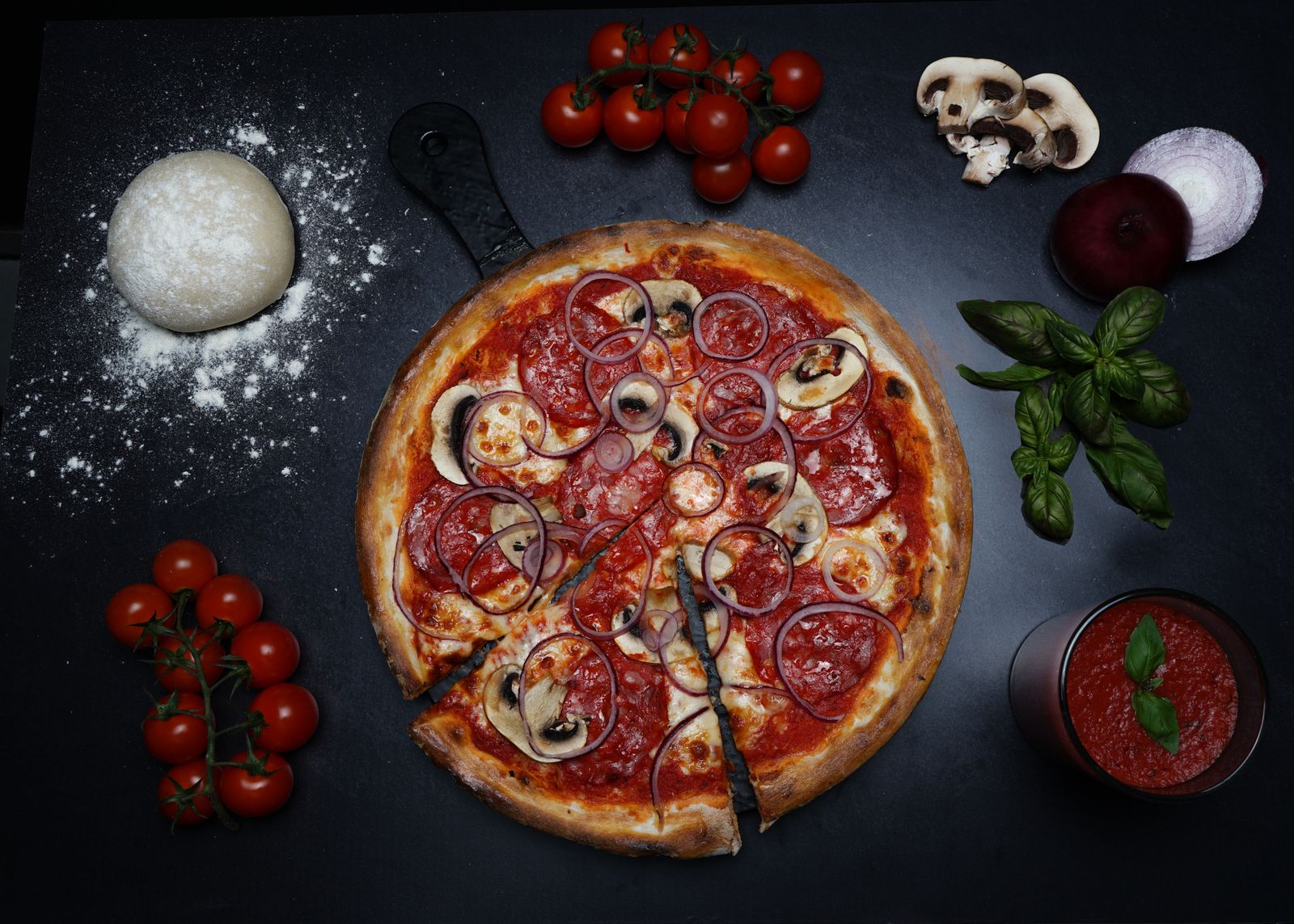 A pizza surrounded by fresh tomatoes, basil and mushrooms
